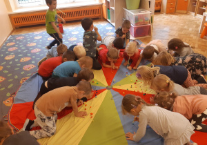 Na dywanie rozłożona jest chusta animacyjna, a na niej położone są pompony. Dzieci trzymają w ustach słomki i próbują z ich pomocą przenieść pompony do pudełek.